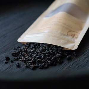 May include: A brown paper bag with a white label is spilling out loose black tea leaves onto a dark wooden surface.