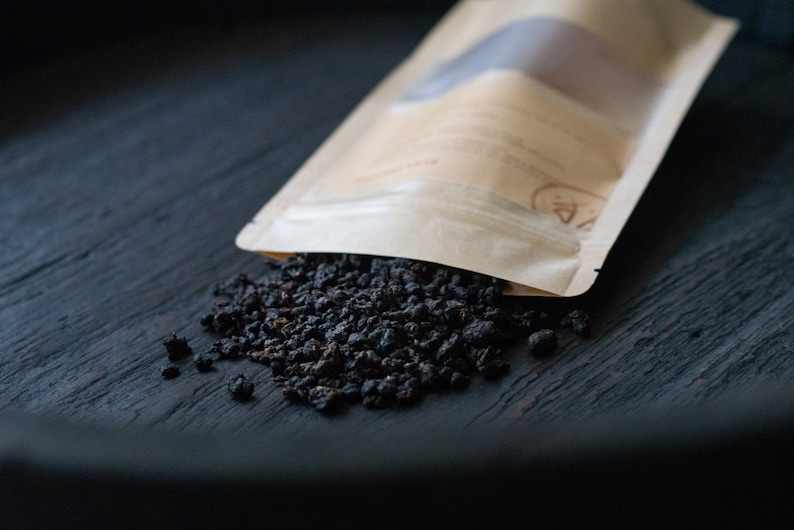 May include: A brown paper bag with a white label is spilling out loose black tea leaves onto a dark wooden surface.