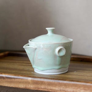Peut inclure: Une théière en céramique avec un vernis vert menthe pâle qui coule vers la base blanche non émaillée. La théière présente un petit bec verseur, une poignée latérale circulaire et un couvercle assorti avec un bouton central, à la texture lisse.