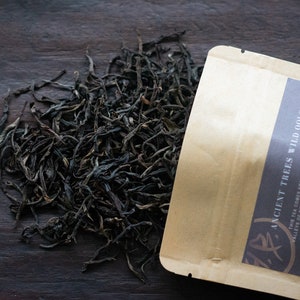 May include: A brown paper bag with the text "ANCIENT TREES WILD OOLONG" printed on it. The bag is open and loose dark tea leaves are spilling out onto a dark wooden surface.