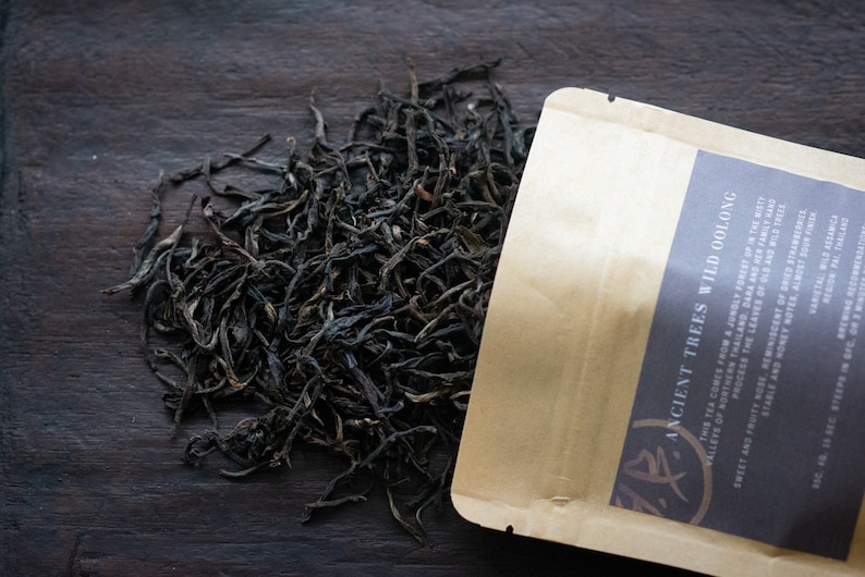 May include: A brown paper bag with the text "ANCIENT TREES WILD OOLONG" printed on it. The bag is open and loose dark tea leaves are spilling out onto a dark wooden surface.
