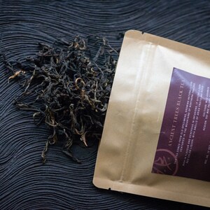 May include: A brown paper bag with a label that reads "Ancient Trees Black Tea". The bag is open and loose black tea leaves are spilling out onto a dark, textured surface.