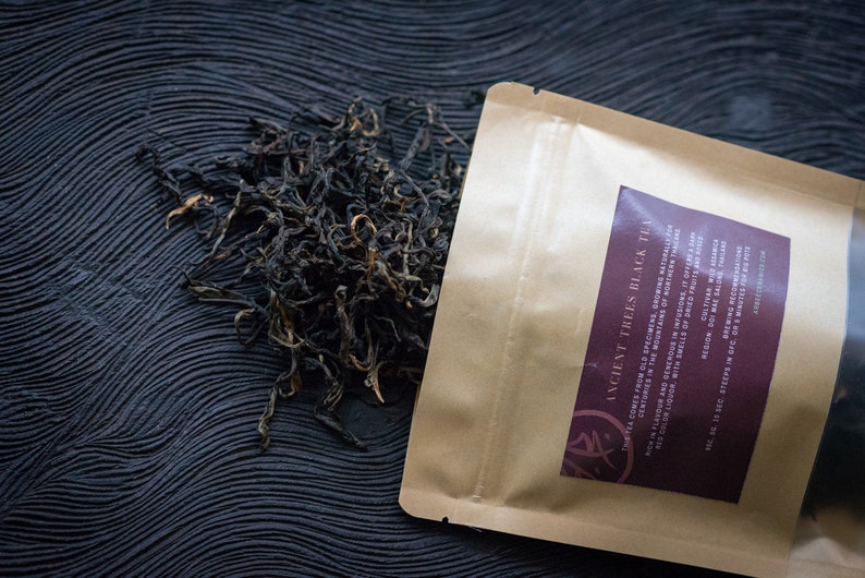May include: A brown paper bag with a label that reads "Ancient Trees Black Tea". The bag is open and loose black tea leaves are spilling out onto a dark, textured surface.