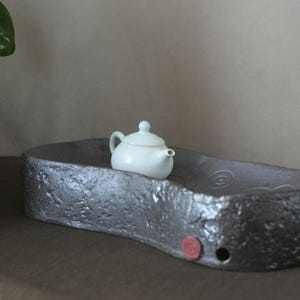 May include: A small, pale celadon-colored ceramic teapot rests on a large, dark gray, textured rectangular tea tray. The tray features a rough, stone-like exterior, a circular red accent, and a small drainage hole on the front side.