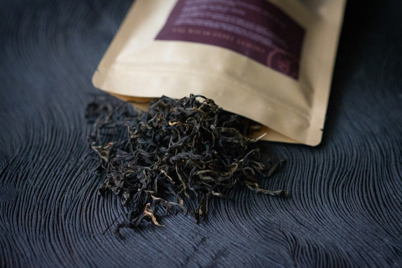 May include: A close-up of a brown paper bag with a label that says "Loose Leaf Black Tea" and a pile of loose leaf black tea on a dark blue surface.