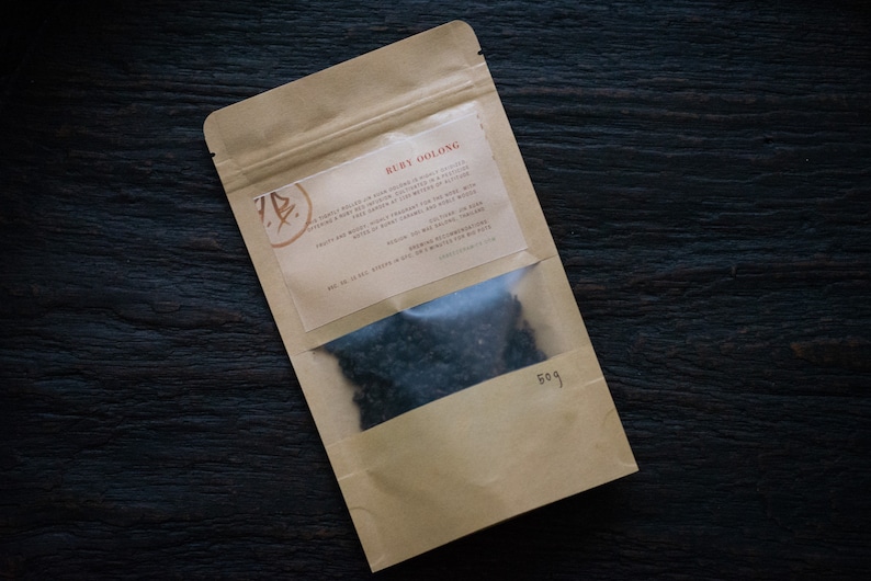May include: A brown paper pouch with a clear window showing loose black tea leaves. The pouch has a label with the text "Ruby Oolong" and a description of the tea.