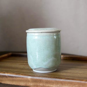 Pode incluir: Um pote cilíndrico de cerâmica com tampa, apresentando um esmalte salpicado de verde menta claro que transita para um acabamento branco liso na base e na borda. A superfície tem uma textura ondulada sutil e a tampa encaixa na abertura.