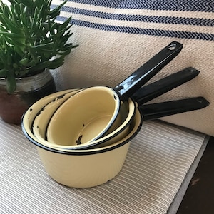 May include: Three vintage enamelware saucepans with black handles. The saucepans are nested together and have a cream-colored exterior with black rims.