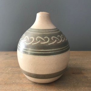 May include: A small, round ceramic vase with a light brown glaze and a dark brown band with a wavy design around the middle.