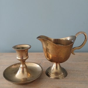 May include: A set of two vintage brass items: a small candlestick with a round base and a creamer with a handle. Both items are made of polished brass and have a warm, golden tone.