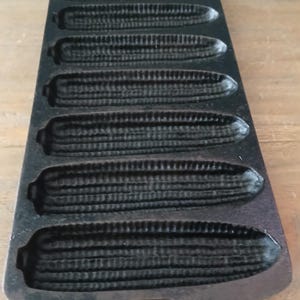 May include: A black cast iron corn stick pan featuring seven elongated, textured molds shaped like ears of corn. The rectangular pan has rounded ends with integrated handles at the top and bottom, designed for baking corn-shaped bread sticks.