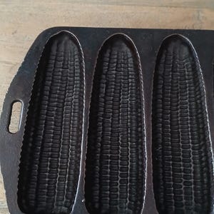 May include: A dark gray cast iron cornbread pan featuring three distinct, elongated, textured molds shaped like ears of corn. The heavy metal pan has a flat, rectangular edge and includes a small, oval-shaped handle opening on the left side.