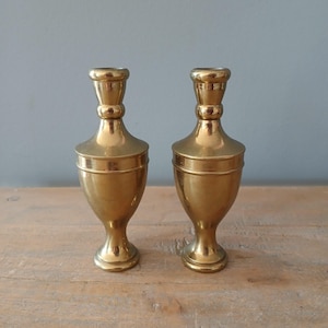 May include: Two gold metal candle holders with a classic design. The holders are shaped like a vase with a round base and a flared top.