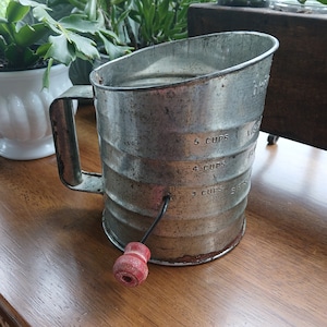May include: A vintage silver-colored metal flour sifter with a red wooden handle. The sifter has markings for 3, 4, and 5 cups.