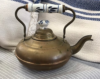Op de afbeelding: Een vintage koperen waterkoker met een blauw en wit porseleinen handvat en een knop voor de deksel. De waterkoker heeft een rond lichaam en een tuit aan de zijkant.