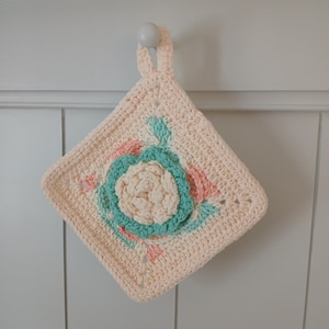 May include: A square, cream-colored crochet potholder with a loop for hanging. The center features a light beige flower with a teal and pink floral design. The potholder is hanging on a white wall.