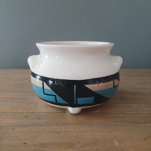 May include: A small, white ceramic pot with two side handles and three small feet. The pot features a band of geometric designs in black, teal, and tan around the middle. The pot is likely used for decorative purposes.