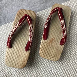 May include: A pair of traditional Japanese wooden sandals, known as geta, with red and white patterned straps. The sandals have a light brown wood finish and a textured surface.