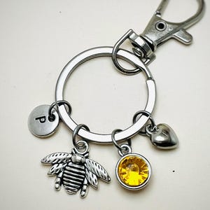May include: Silver-toned keychain with a round key ring and a lobster clasp. Charms include a bee, a heart, a yellow gemstone, and a disc with the letter "P". The keychain is designed to hold keys or be attached to a bag.