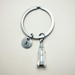 May include: Silver-coloured keyring with a rocket charm and a circular tag bearing the letter "X". The rocket charm has detailed design elements. The keyring is also silver-coloured.