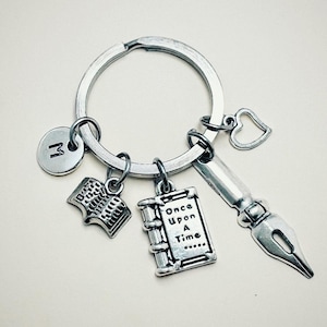 Puede incluir: Llavero de plata con abalorios, incluyendo un libro, una pluma, un corazón y un abalorio con la letra M. El abalorio del libro tiene el texto "Once Upon A Time".