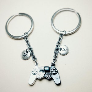 May include: A silver keychain with two key rings connected by a chain. The chain has two small silver charms with the letters 'A' and 'V' on them. The keychain also has a charm in the shape of a video game controller, half black and half white.