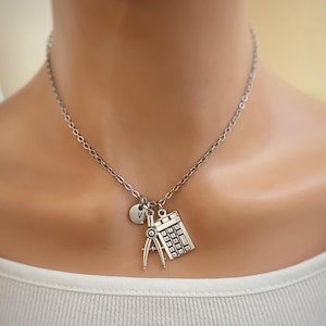 Può includere: Collana color argento con ciondoli a forma di calcolatrice, compasso e un ciondolo circolare con la lettera "V". La collana è indossata da una persona con una maglia bianca.