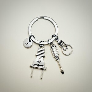 May include: Silver keychain with a lightbulb, screwdriver, and electrical plug charm. The plug charm has a lightning bolt and the letter 'J' is on a round charm.