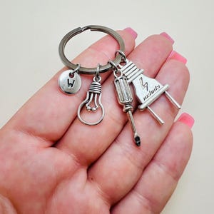 May include: Silver keychain with a lightbulb, screwdriver, and electrical plug charm. The keychain also has a small charm with the letter "W" on it.