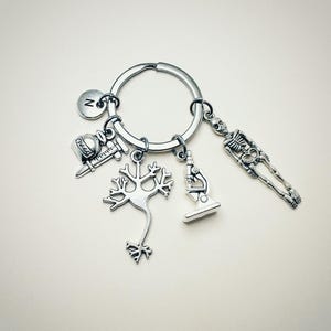 Op de afbeelding: Zilveren sleutelhanger met verschillende bedels, waaronder een skelet, een microscoop, een neuron, een spuit en een flesje. De sleutelhanger heeft ook een bedel met de letter 'N'.