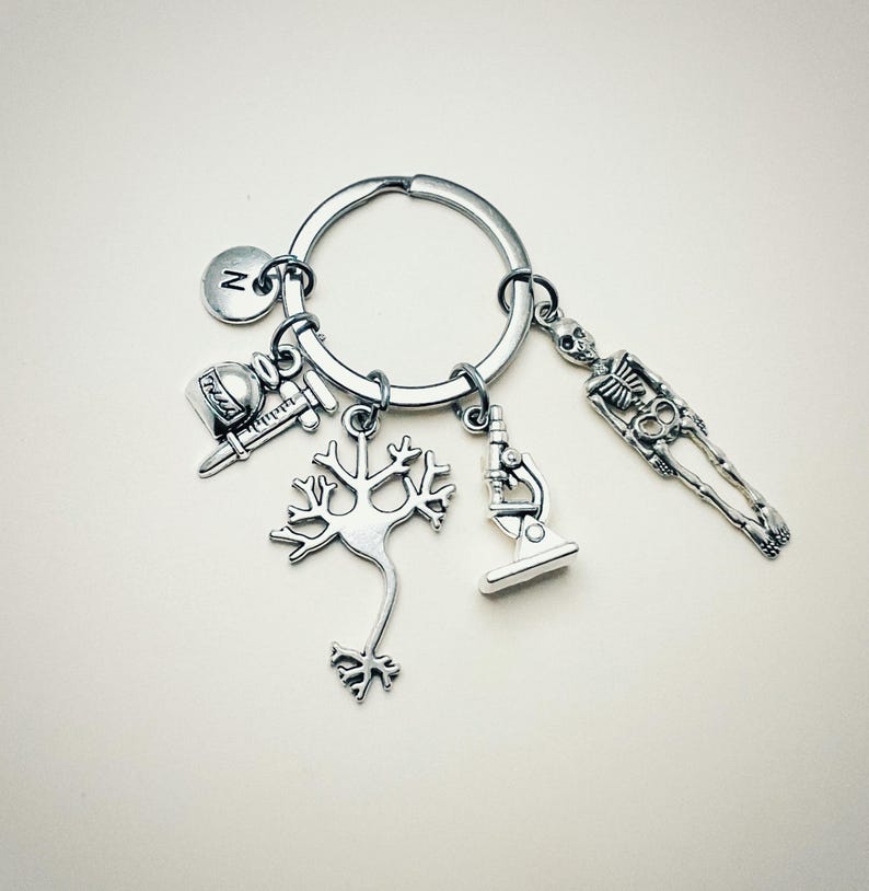 Op de afbeelding: Zilveren sleutelhanger met verschillende bedels, waaronder een skelet, een microscoop, een neuron, een spuit en een flesje. De sleutelhanger heeft ook een bedel met de letter 'N'.
