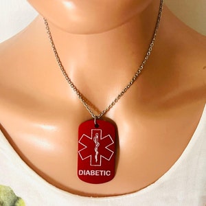 Può includere: Ciondolo a targhetta rosso su una catena d'argento. La targhetta presenta un simbolo di allerta medica bianco e la parola "DIABETIC" in bianco. Il ciondolo è rettangolare con angoli arrotondati.