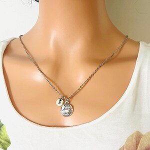 Puede incluir: Collar plateado con un colgante de globo y un pequeño dije circular con la letra "B". El collar se muestra sobre una blusa blanca, resaltando el diseño y los detalles de la joya.