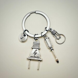 May include: Silver keychain with a lightbulb, screwdriver, and electrical plug charm. The plug charm has a lightning bolt symbol. The keychain also has a round charm with the letter J.