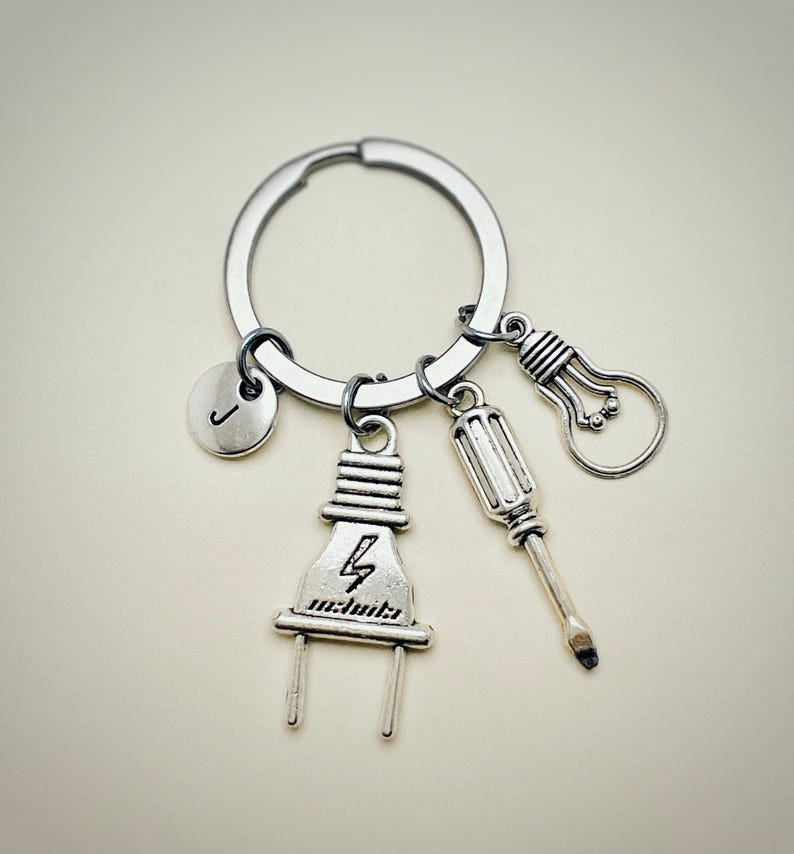 May include: Silver keychain with a lightbulb, screwdriver, and electrical plug charm. The plug charm has a lightning bolt symbol. The keychain also has a round charm with the letter J.
