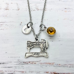 Può includere: Una collana d'argento con un ciondolo a forma di macchina da cucire vintage, un piccolo ciondolo rotondo con la lettera "E" e un ciondolo di strass giallo.