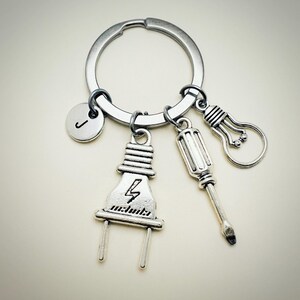 May include: Silver keychain with a lightbulb, screwdriver, and electrical plug charm. The plug charm has a lightning bolt design. The keychain also has a silver initial charm with the letter "J" on it.
