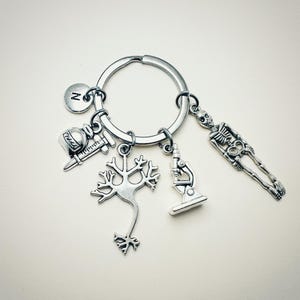 Op de afbeelding: Zilveren sleutelhanger met verschillende bedels, waaronder een skelet, een microscoop, een neuron, een flesje met een spuit en een bedel met de letter 'N'.