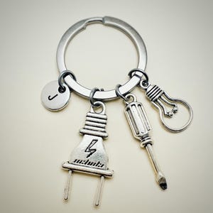 May include: Silver keychain with a lightbulb, screwdriver, and electrical plug charm. The keychain also has a silver charm with the letter J.