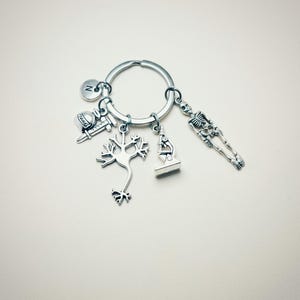 Op de afbeelding: Zilveren sleutelhanger met een verscheidenheid aan bedels, waaronder een neuron, een microscoop, een skelet, een spuit en een afstudeerhoed. De sleutelhanger heeft ook een kleine bedel met de letter "N" erop.