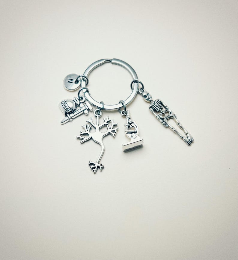 Op de afbeelding: Zilveren sleutelhanger met een verscheidenheid aan bedels, waaronder een neuron, een microscoop, een skelet, een spuit en een afstudeerhoed. De sleutelhanger heeft ook een kleine bedel met de letter "N" erop.