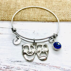 Könnte beinhalten: Verstellbares Armreif-Armband in Silber mit einem runden Anhänger mit dem Buchstaben "P", Komödien- und Tragödienmasken und einem blauen Edelstein. Das Armband wird auf einer weißen Holzoberfläche präsentiert.