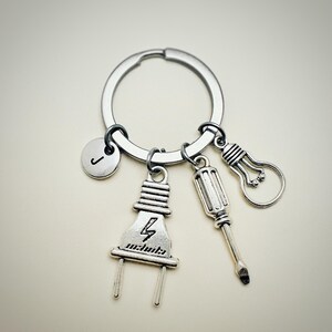 May include: Silver keychain with a lightbulb, screwdriver, and electrical plug charm. The plug charm has a lightning bolt symbol. The keychain also has a round charm with the letter J.