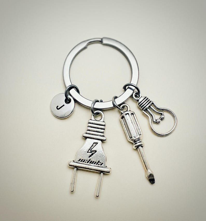 May include: Silver keychain with a lightbulb, screwdriver, and electrical plug charm. The plug charm has a lightning bolt symbol. The keychain also has a round charm with the letter J.
