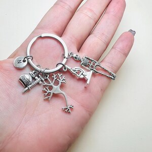 Op de afbeelding: Zilveren sleutelhanger met verschillende bedels, waaronder een skelet, een microscoop, een neuron, een flesje met een spuit en een bedel met de letter 'N'.