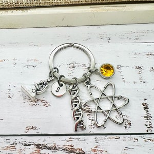 Puede incluir: Llavero plateado con temática científica con un microscopio, una cadena de ADN, un símbolo de átomo y un dije de piedra preciosa amarilla. Incluye un dije circular con la letra "S". El llavero está unido a un anillo plateado.