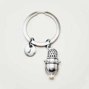 Könnte beinhalten: Silberfarbener Schlüsselanhänger mit einem Mikrofon-Anhänger und einem runden Anhänger mit dem Buchstaben "J". Der Mikrofon-Anhänger hat ein Vintage-Design mit detailliertem Grill. Der Schlüsselanhänger ist an einem silbernen Ring befestigt.