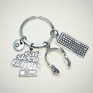 Può includere: Portachiavi d'argento con un ciondolo che dice "mangia dormi gioca" e ha un design di controller per videogiochi. Il portachiavi ha anche ciondoli di una tastiera, cuffie e una lettera O.