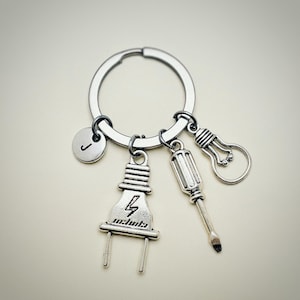 May include: Silver keychain with a letter J charm, a lightbulb charm, a screwdriver charm, and a plug charm. The plug charm has a lightning bolt and the word "electricity" on it.