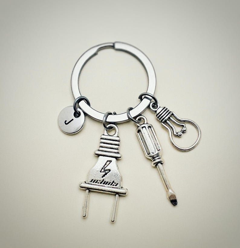 May include: Silver keychain with a letter J charm, a lightbulb charm, a screwdriver charm, and a plug charm. The plug charm has a lightning bolt and the word "electricity" on it.